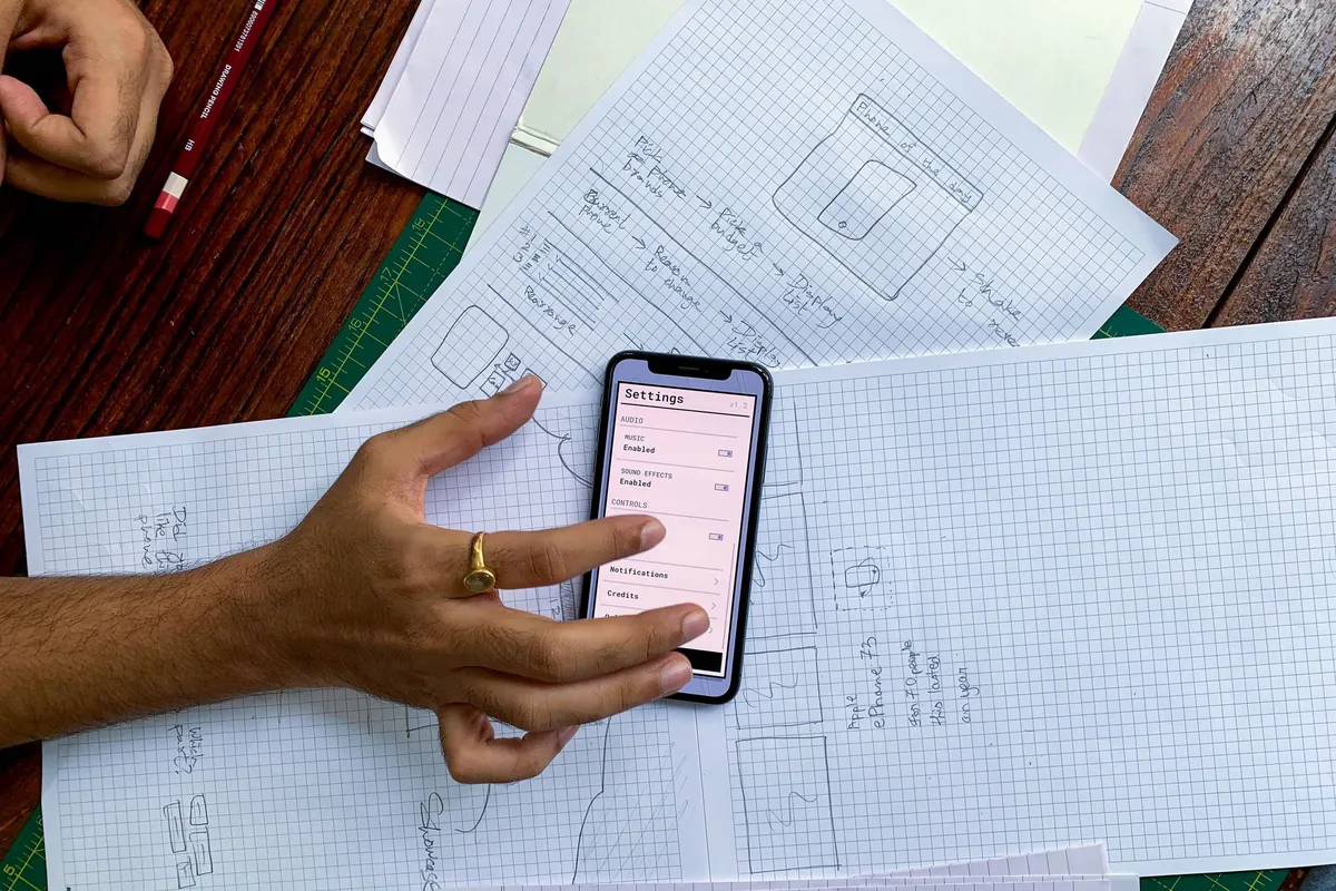 Hand holding a smartphone over paper wireframe sketches of mobile app screens