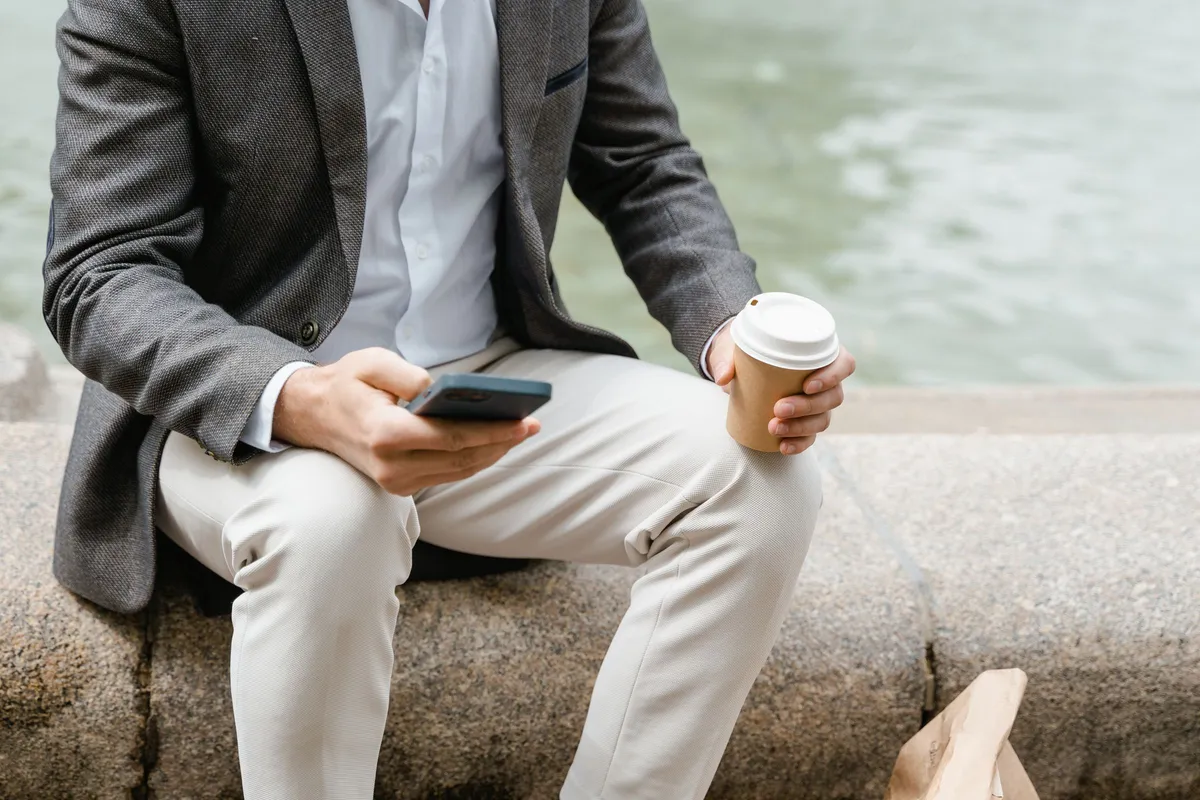 Man in a blazer seated outdoors holding a smartphone and a coffee cup, face out of frame