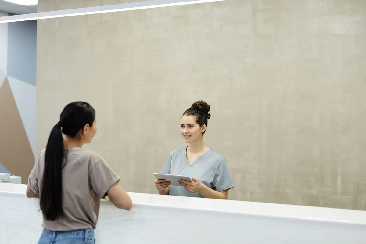 Patient seen from behind at a clinic reception desk while the receptionist checks a booking on a tablet