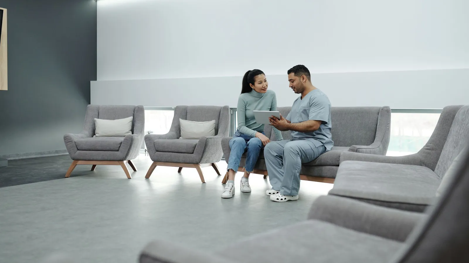 Clinic waiting area with grey armchairs where a staff member shows a patient details on a tablet