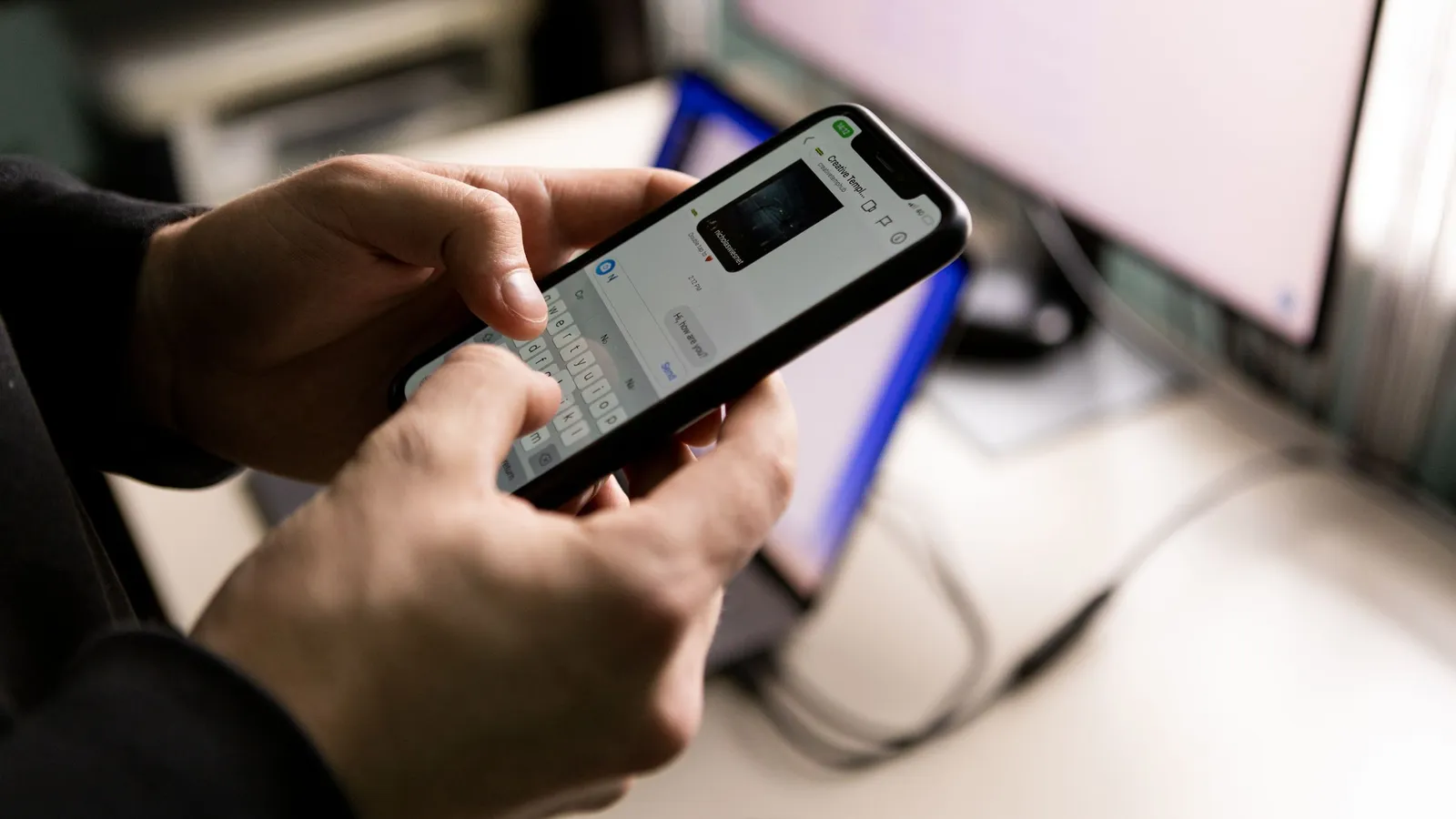 Close-up of hands holding a smartphone with a chat conversation open, a computer screen in the background