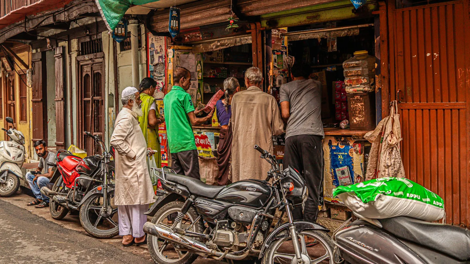 Customers gathered at a busy neighbourhood shop on an Indian street with motorcycles parked outside