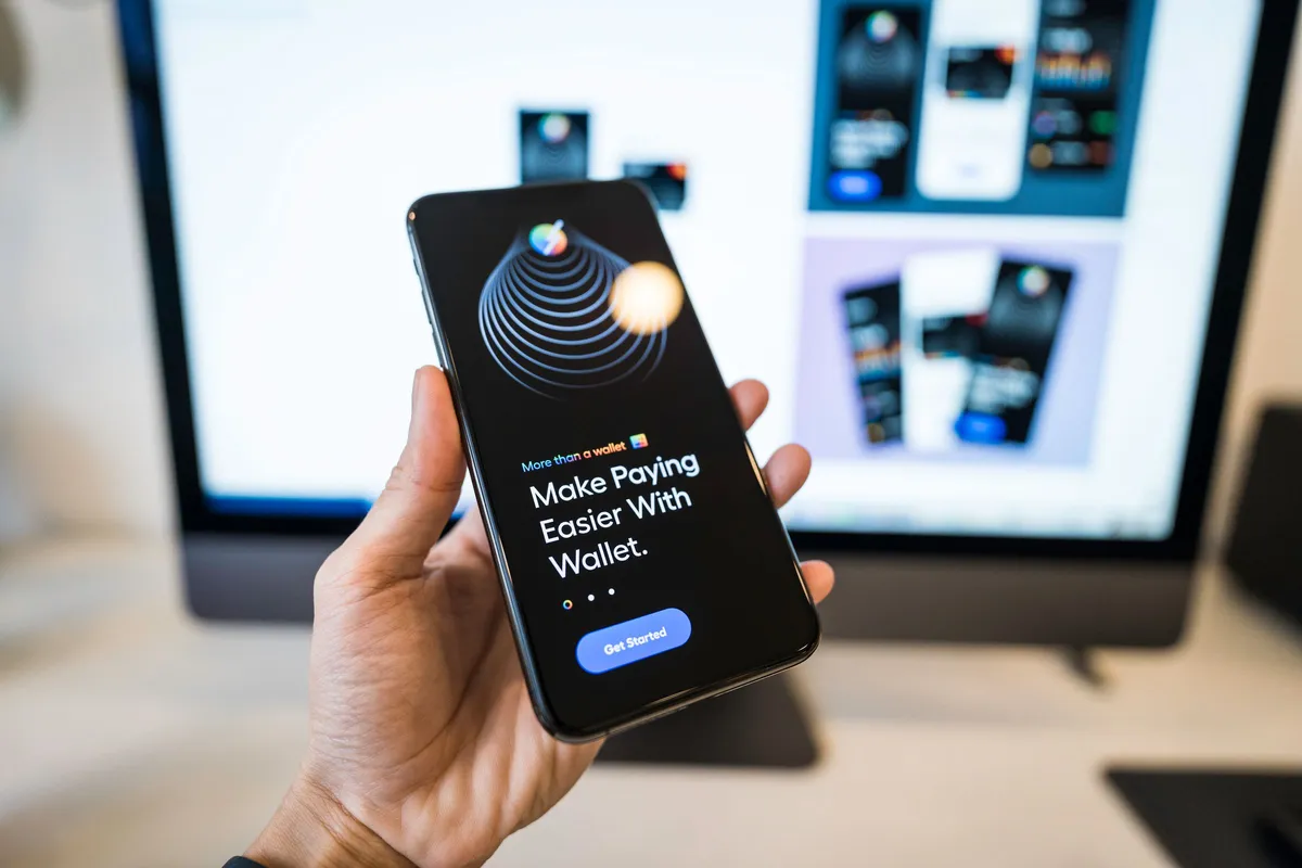 Hand holding a smartphone displaying a wallet app design in front of monitors showing UI layouts