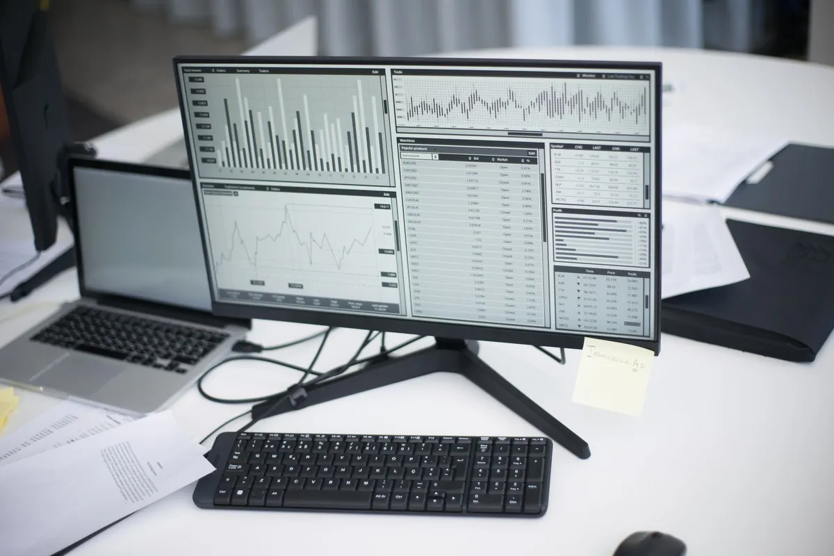 Office desk with a large monitor and a laptop showing spreadsheet reports and charts