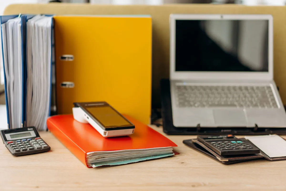 Office desk with ring binders, folders, a calculator, card machine and a laptop