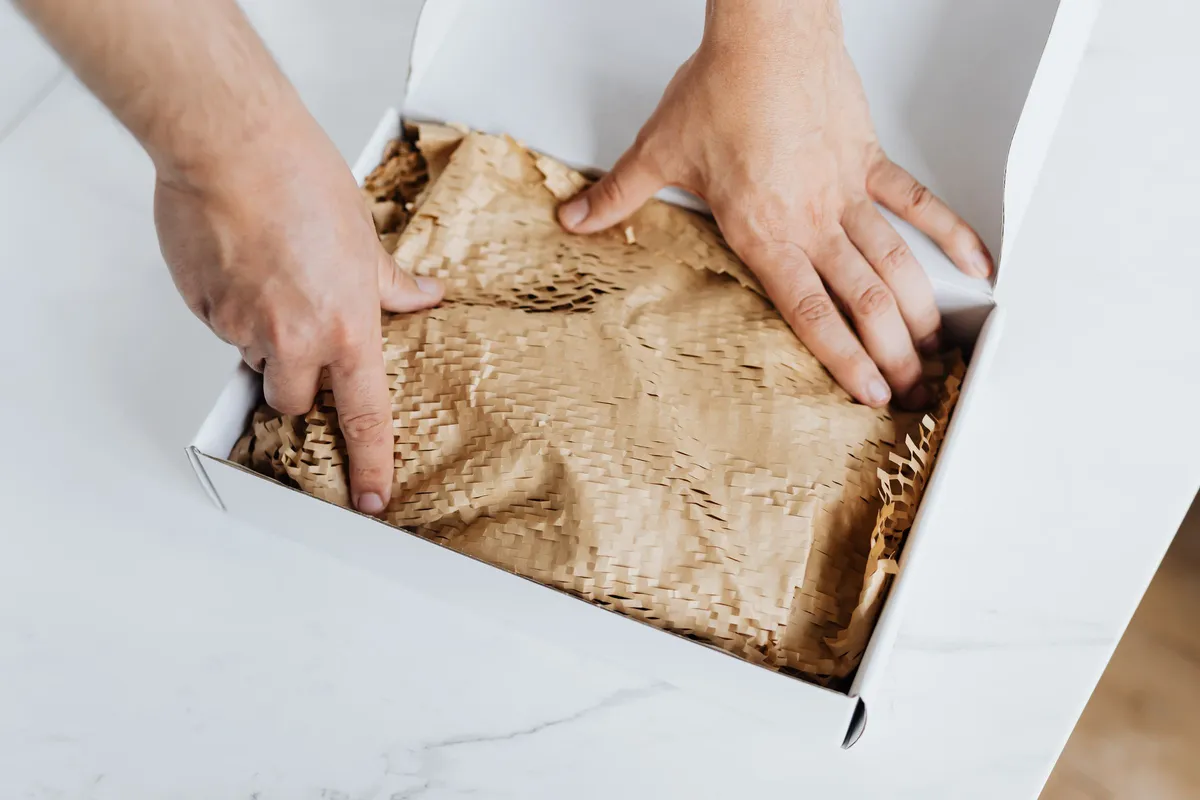 Hands placing paper padding into a cardboard box while packing a product for shipment