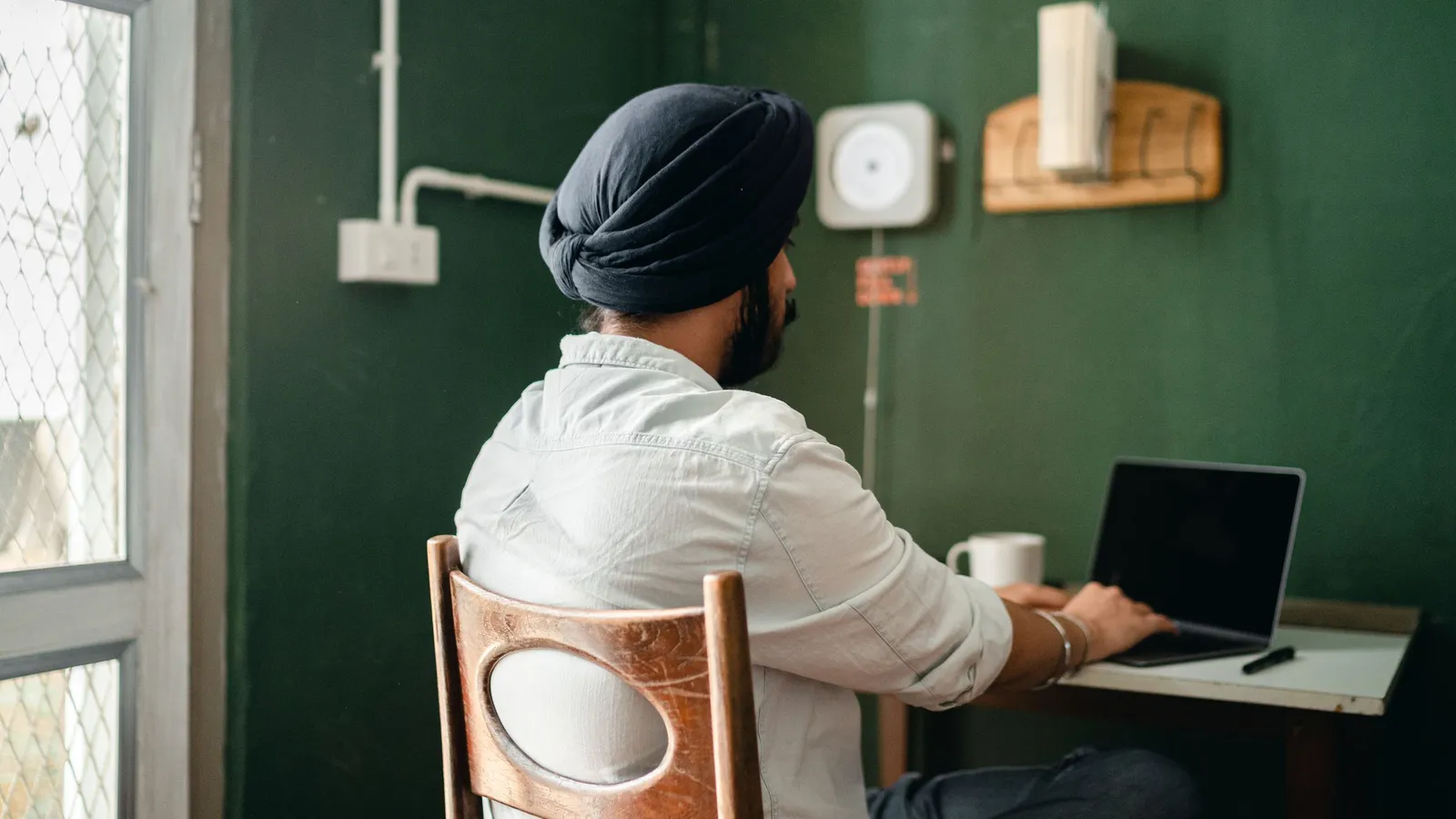 Indian business owner seen from behind, working on a laptop at a small desk in a home office