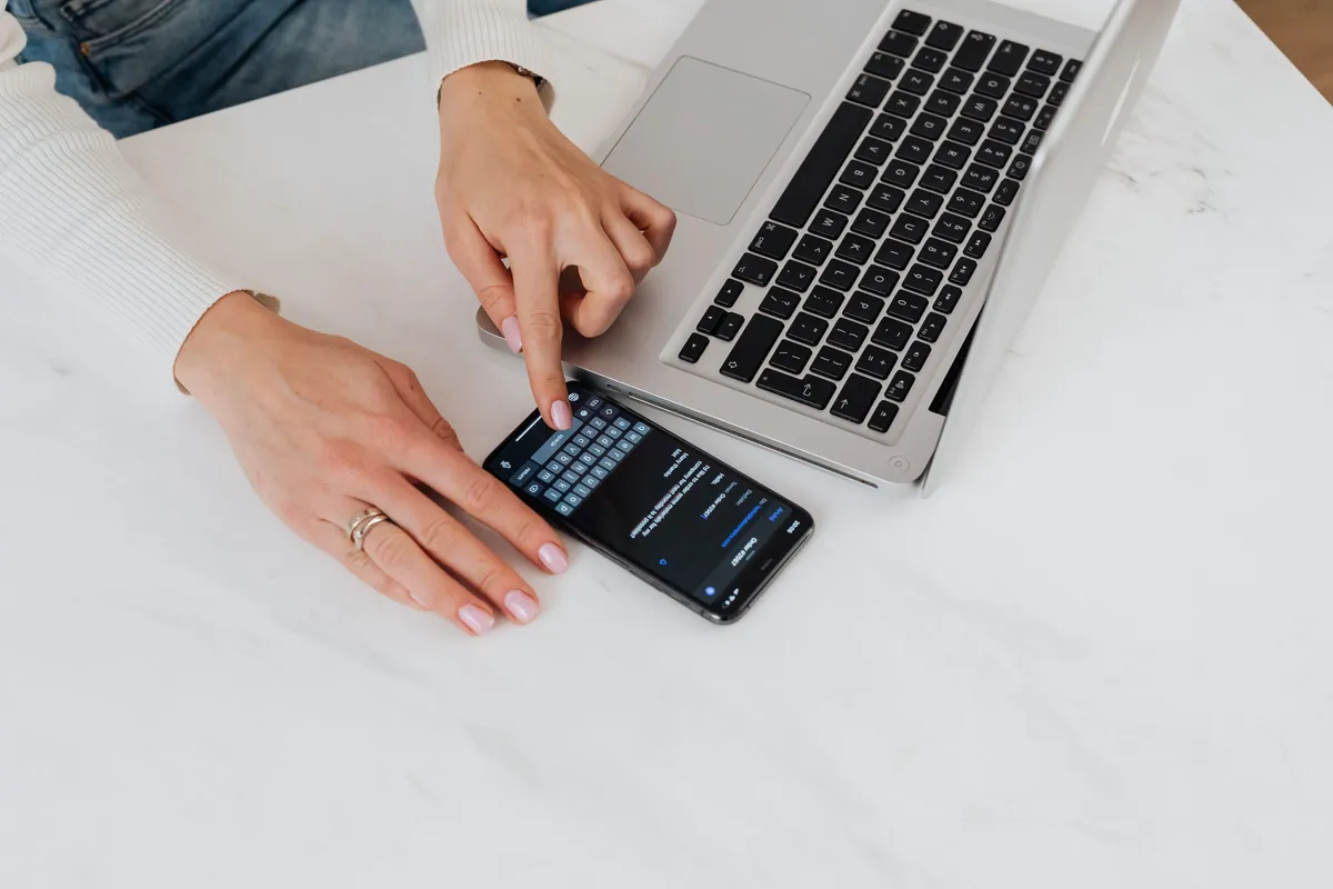 Top view of hands typing a message on a smartphone next to an open laptop on a white desk