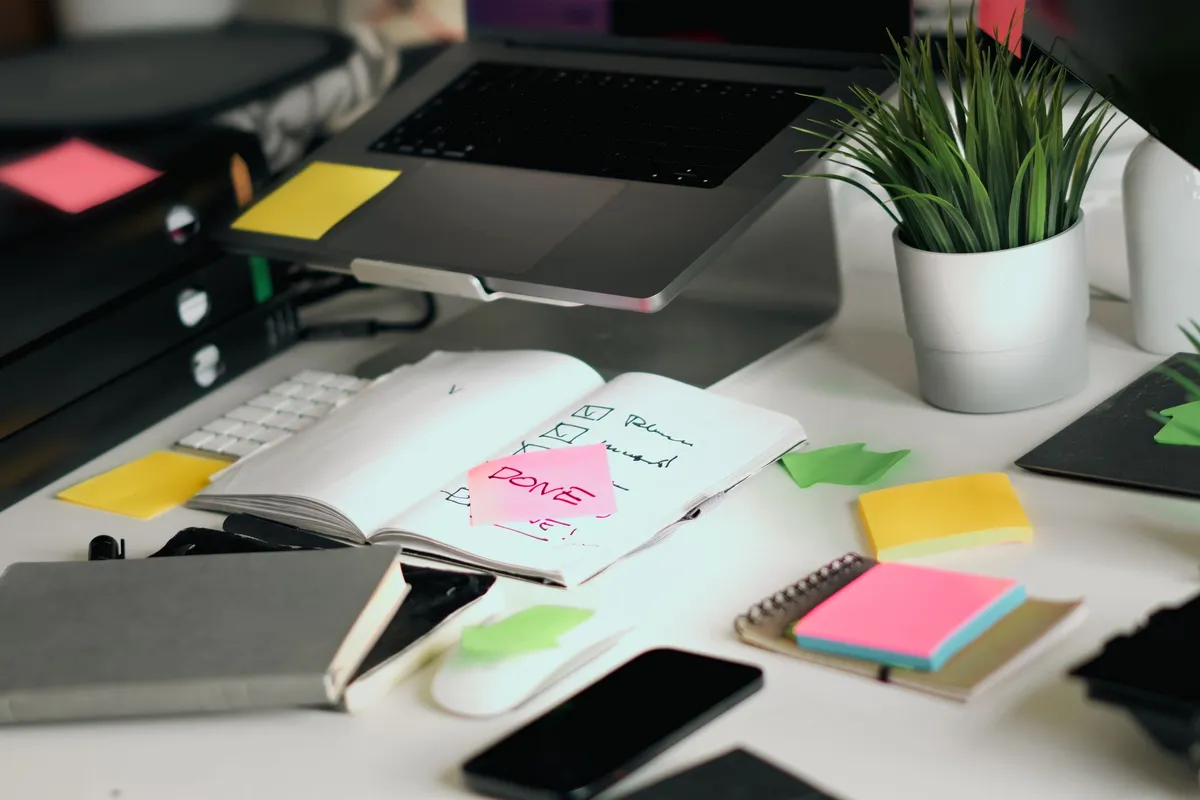 Desk with a notebook, colourful sticky notes, a plant and a laptop used for planning tasks